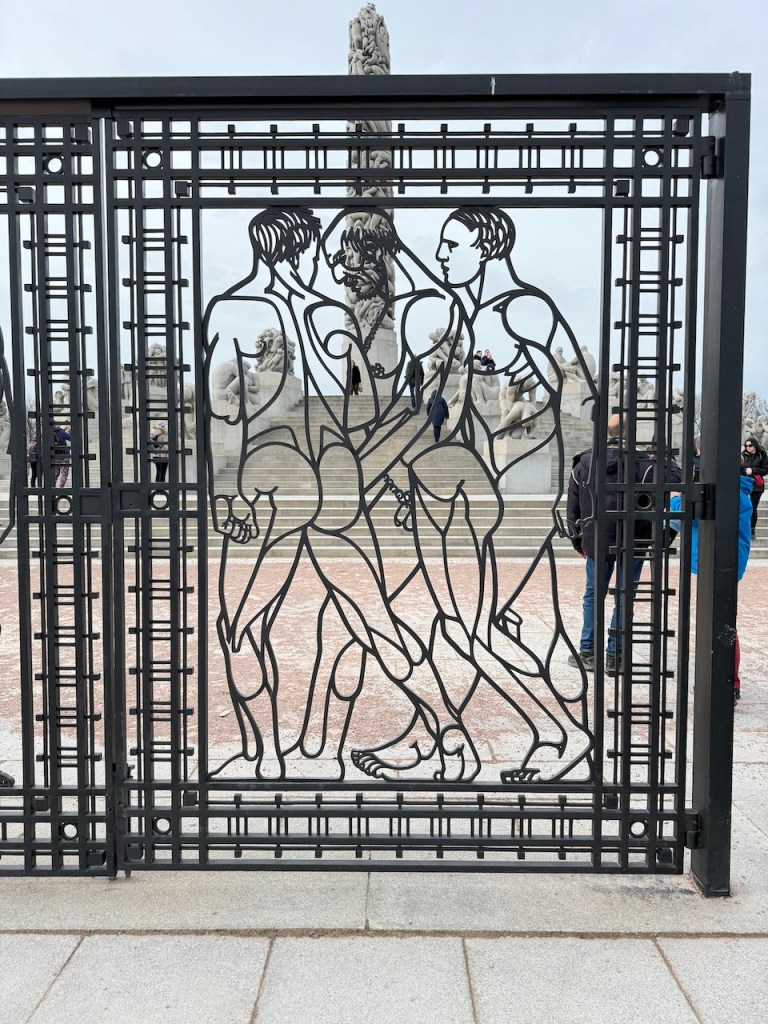 A decorative metal gate featuring a stylized depiction of three human figures, located in a sculpture park.