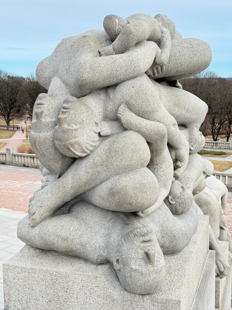 A close-up of a intricate granite sculpture featuring a mass of intertwined human figures, capturing various poses and expressions, set against a serene park background.