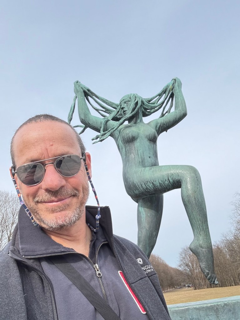 A person posing for a selfie in front of a large, bronze sculpture of a woman with flowing hair, set in a park.