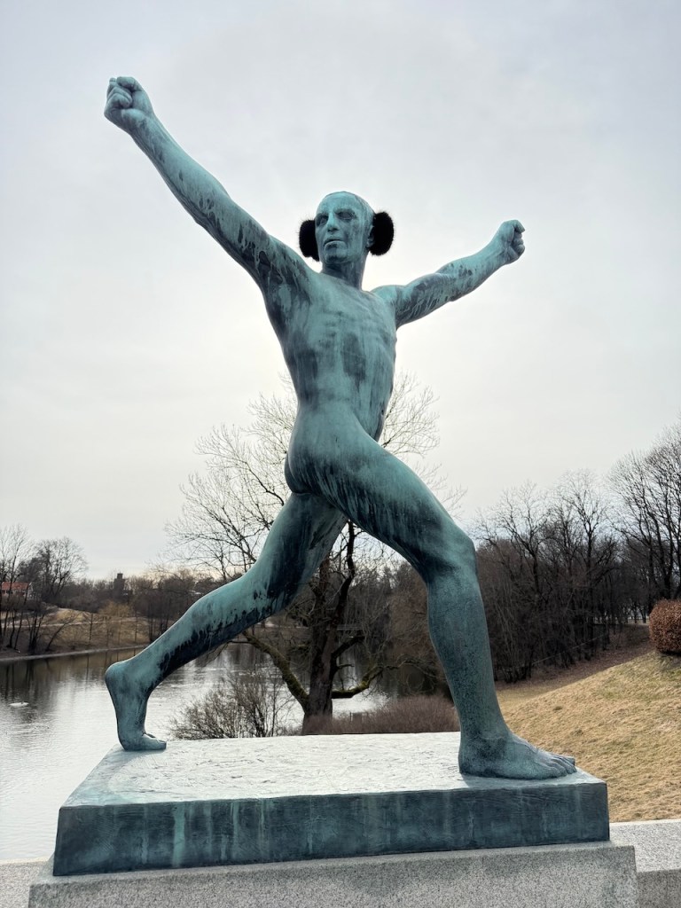 A bronze statue of a muscular athlete in motion, raising one arm triumphantly, with a serene park background featuring trees and a lake.