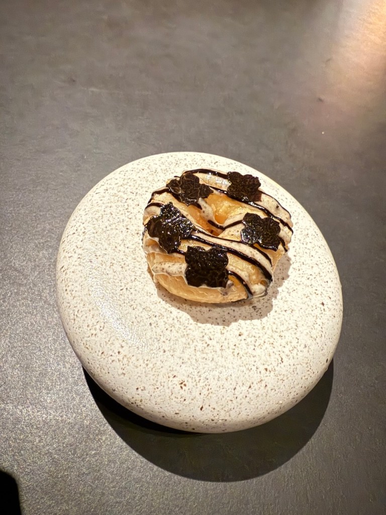 A gourmet dessert featuring a pastry topped with dark chocolate drizzle and decorative pieces on a speckled white plate.