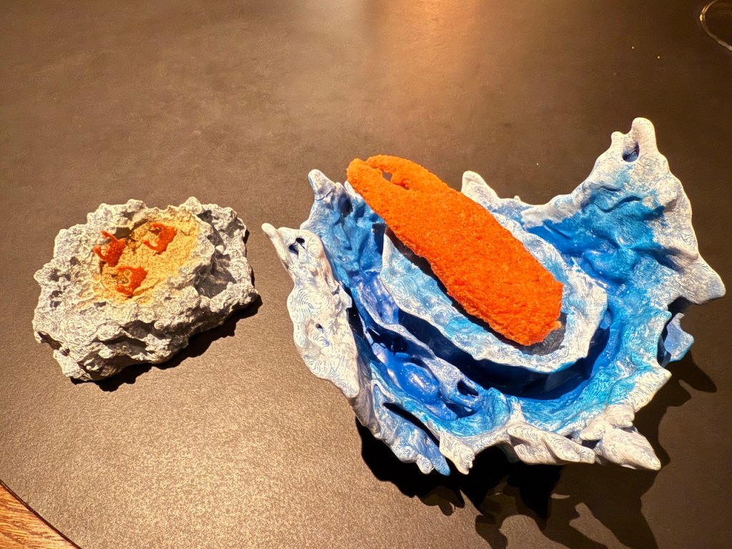 A colorful culinary presentation featuring two artistic plates: one with a textured blue wave design holding a bright orange element resembling seafood, and another with a rocky texture displaying small orange details. Both plates are set against a dark surface.