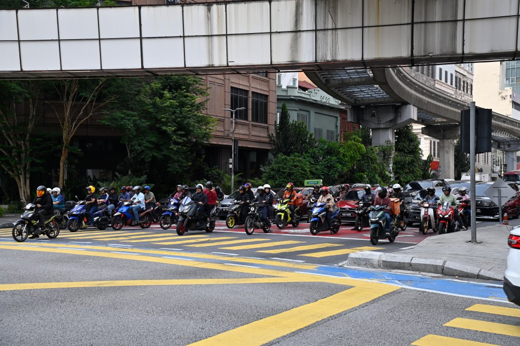 A busy intersection in Kuala Lumpur showing numerous motorcycles waiting at a traffic light, with a monorail track above and buildings in the background.