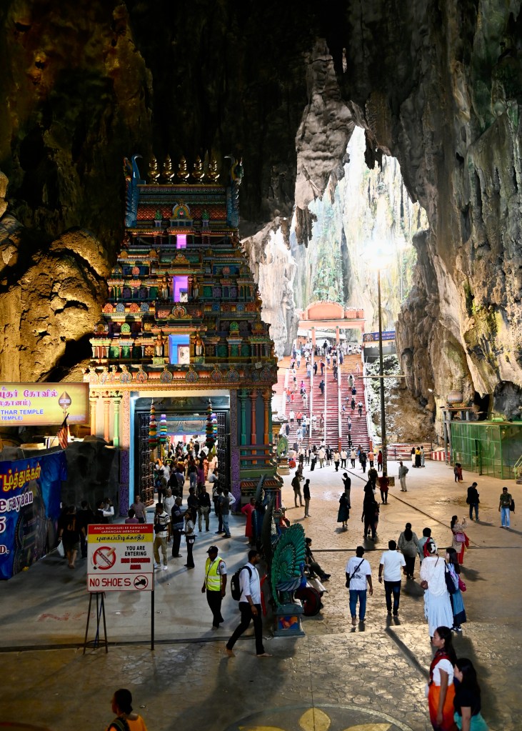 View of the entrance to the Batu Caves, featuring a colorful temple structure and a large crowd of visitors in the cavernous space, with steep stairs leading further into the cave.