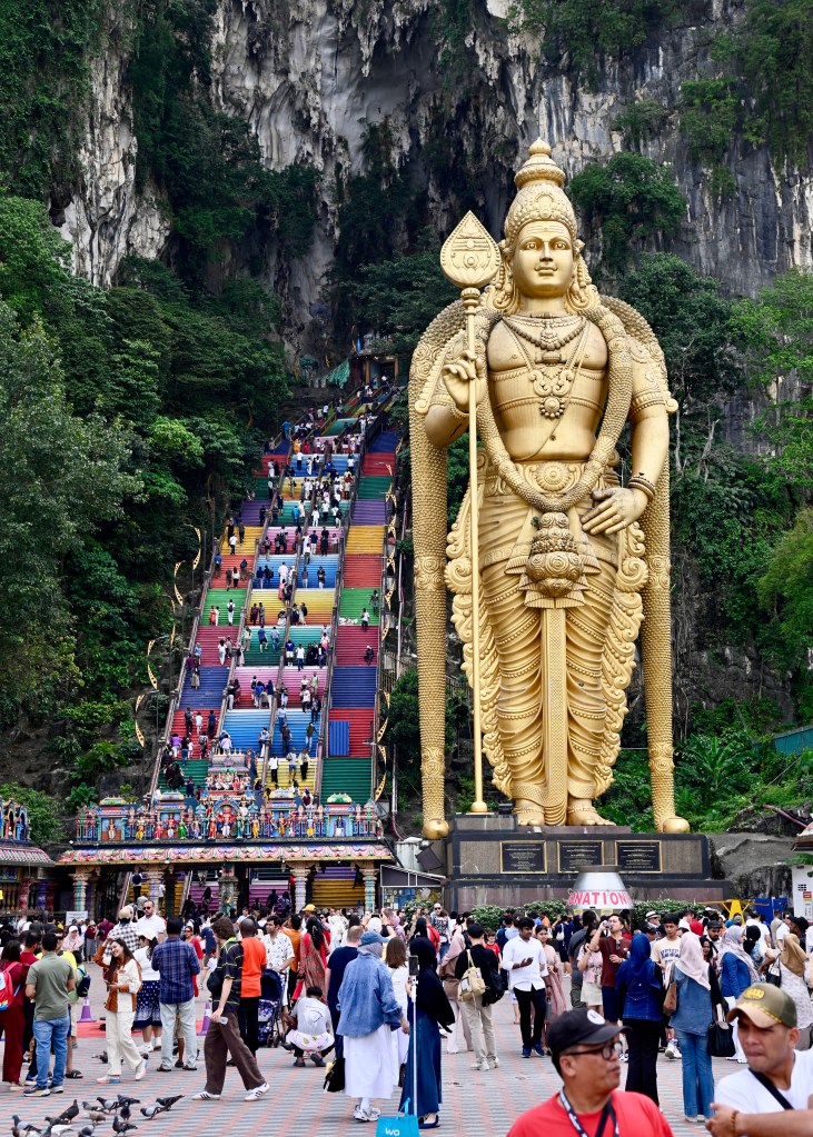 A large golden statue of the Hindu god Murugan stands at the Batu Caves entrance, surrounded by visitors. Colorful steps ascend the limestone mountain in the background.
