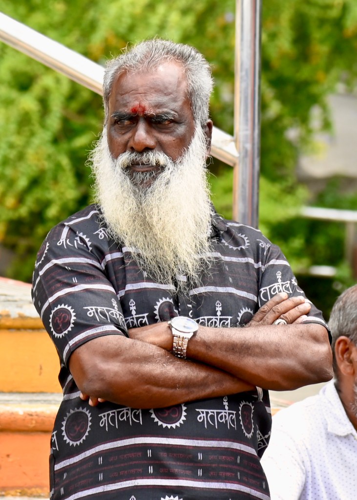 A man with a long white beard wearing a black patterned shirt, standing with arms crossed and a serious expression.