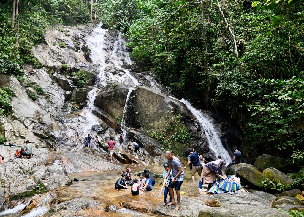 A scenic view of Kanching Waterfalls with people enjoying the natural pool and surroundings, surrounded by lush greenery and rocky terrain.