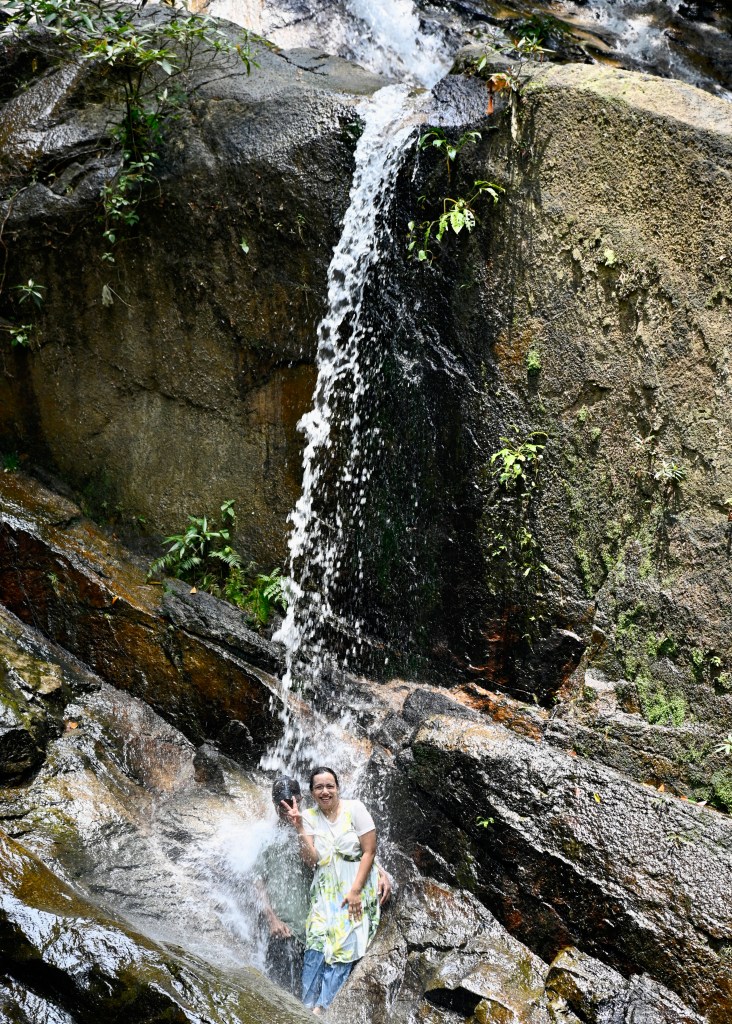 A woman standing under a waterfall at Kanching Waterfalls, with water cascading down rocky terrain surrounded by lush greenery.