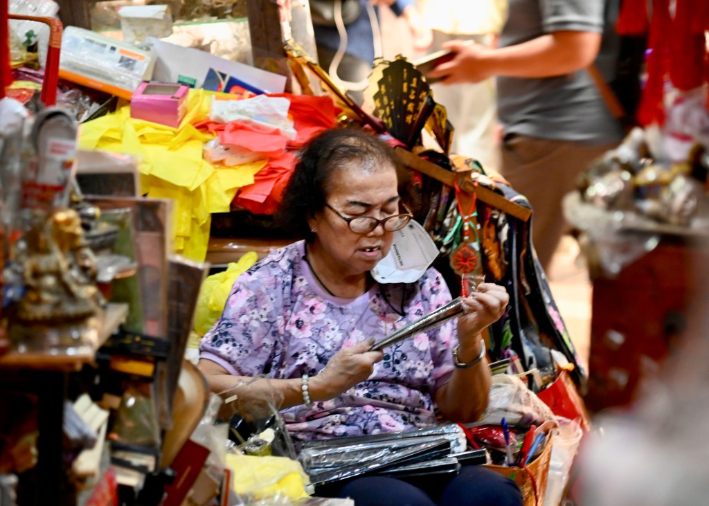 A woman sitting on the floor of a vibrant market, surrounded by colorful items and decorations, intently examining a silver object.