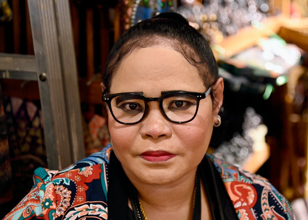 A close-up portrait of a woman with glasses, wearing a colorful patterned shirt, set against a backdrop of various art and craft items.