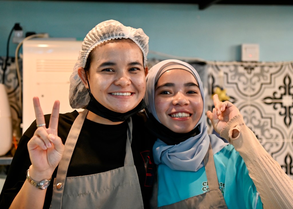 Two smiling women posing together, both wearing aprons. One woman has a hairnet and is making a peace sign with her left hand, while the other woman is smiling and making a peace sign with her right hand. The background features a patterned wall.