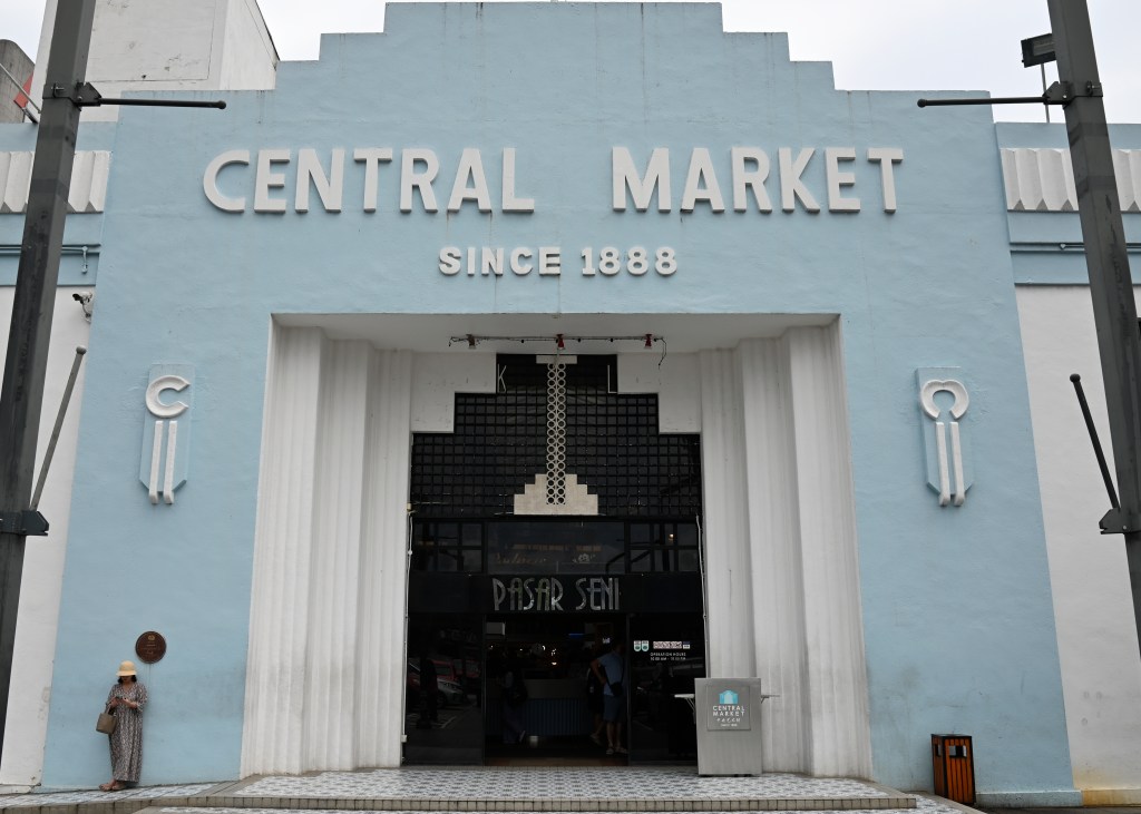 Facade of the Central Market in Kuala Lumpur, featuring the entrance with the words 'CENTRAL MARKET' and 'SINCE 1888', a pastel blue exterior, and a person standing nearby.