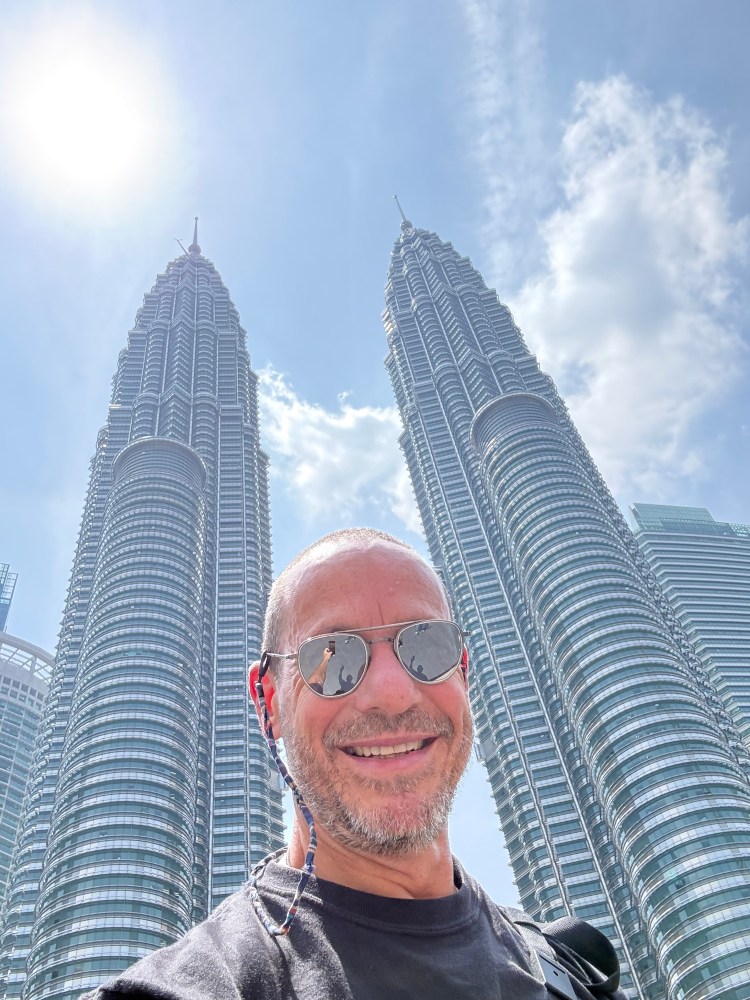 Selfie in front of the Petronas Towers in Kuala Lumpur, showcasing the tall twin skyscrapers against a blue sky.