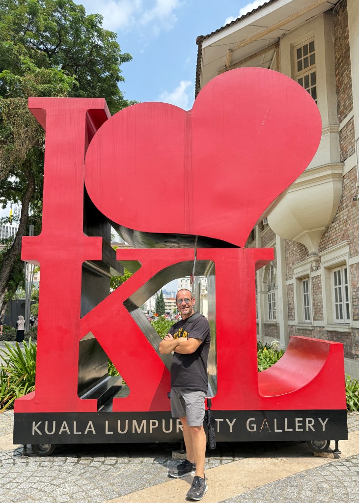 A person standing beside a large 'I ❤️ KL' sculpture in Kuala Lumpur, with a city gallery in the background.