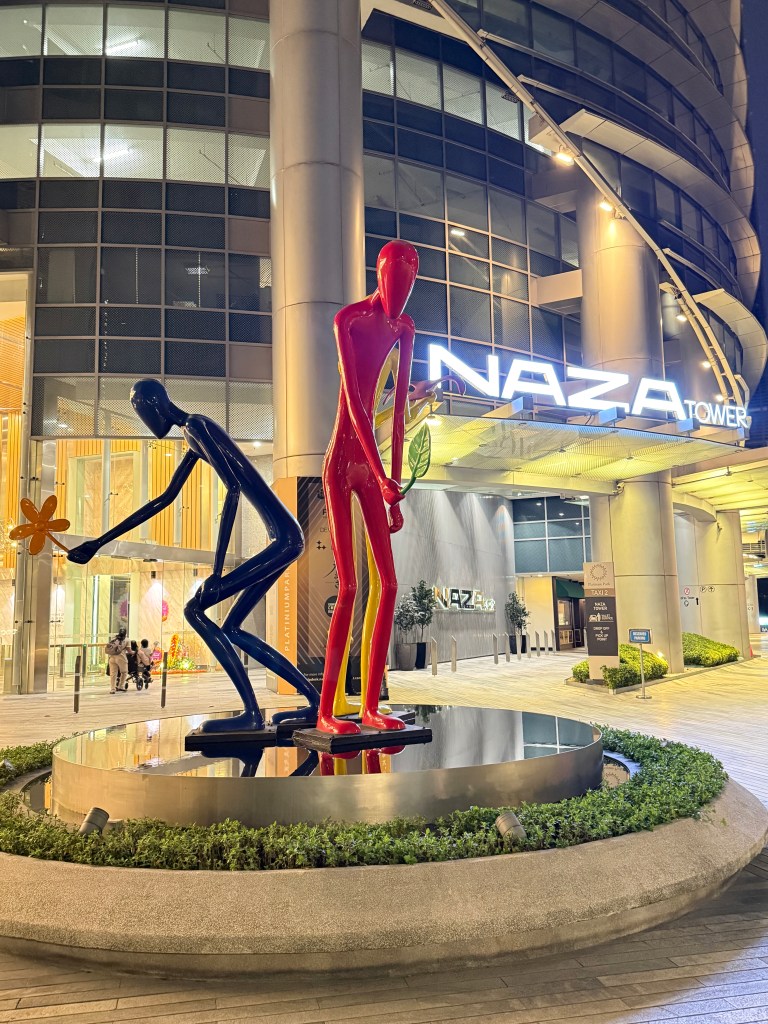 Colorful art sculptures of two stick figures, one red and one blue, near the entrance of Naza Tower in Kuala Lumpur, with the building's signage visible in the background.