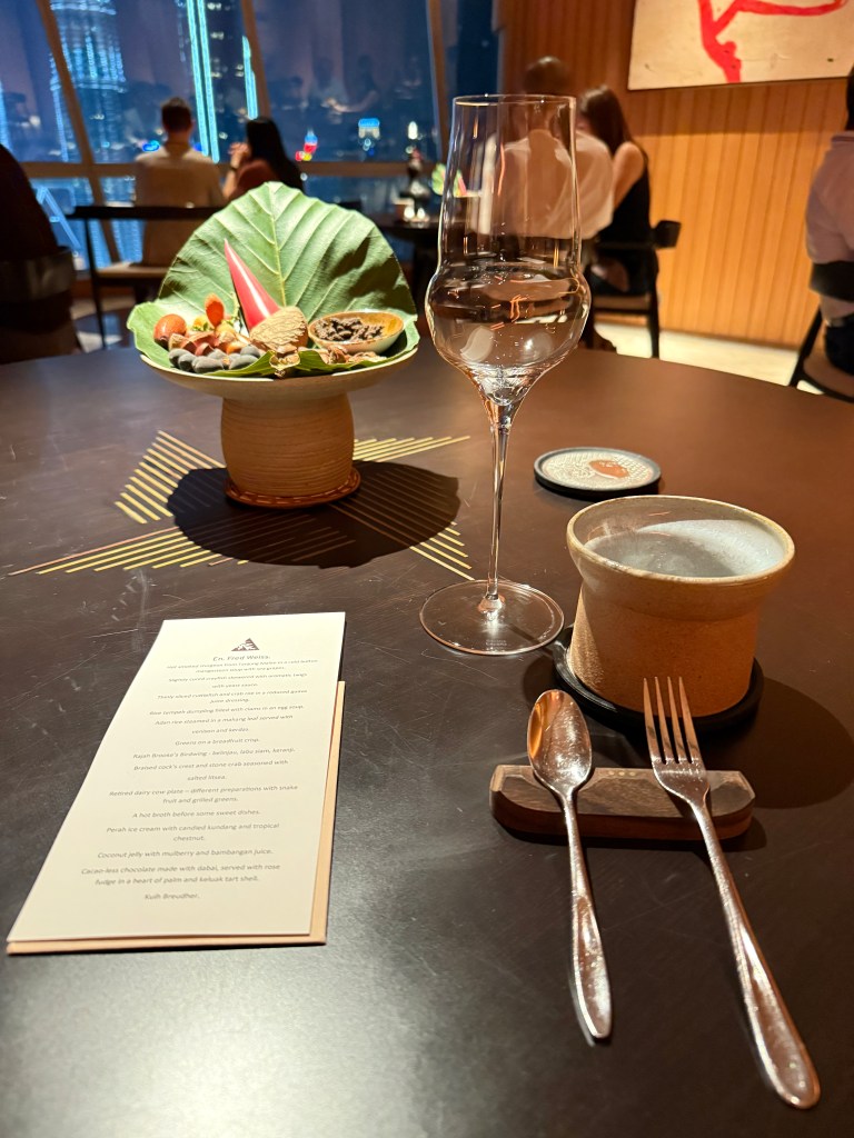 A beautifully arranged dining table at a Michelin-starred restaurant in Kuala Lumpur, featuring a decorative plate with assorted appetizers, a glass of water, and a menu.