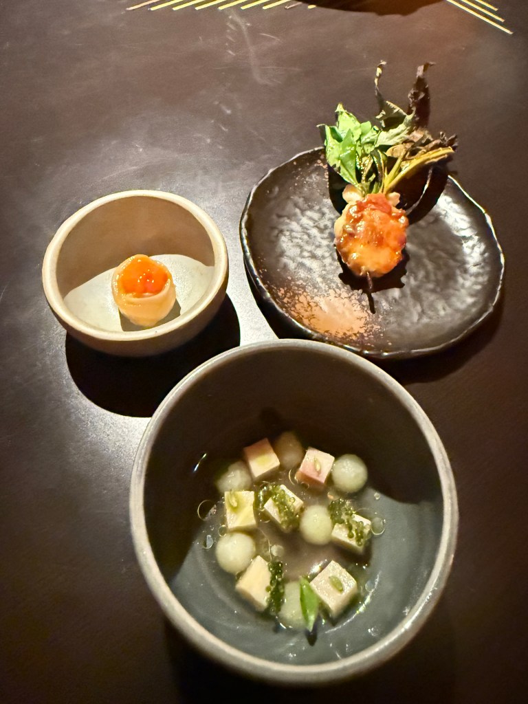A beautifully presented three-course dish featuring a variety of small servings, including a round item with a bright orange topping, a delicate shrimp garnished with greens, and a bowl with cubes and spheres in a clear broth.