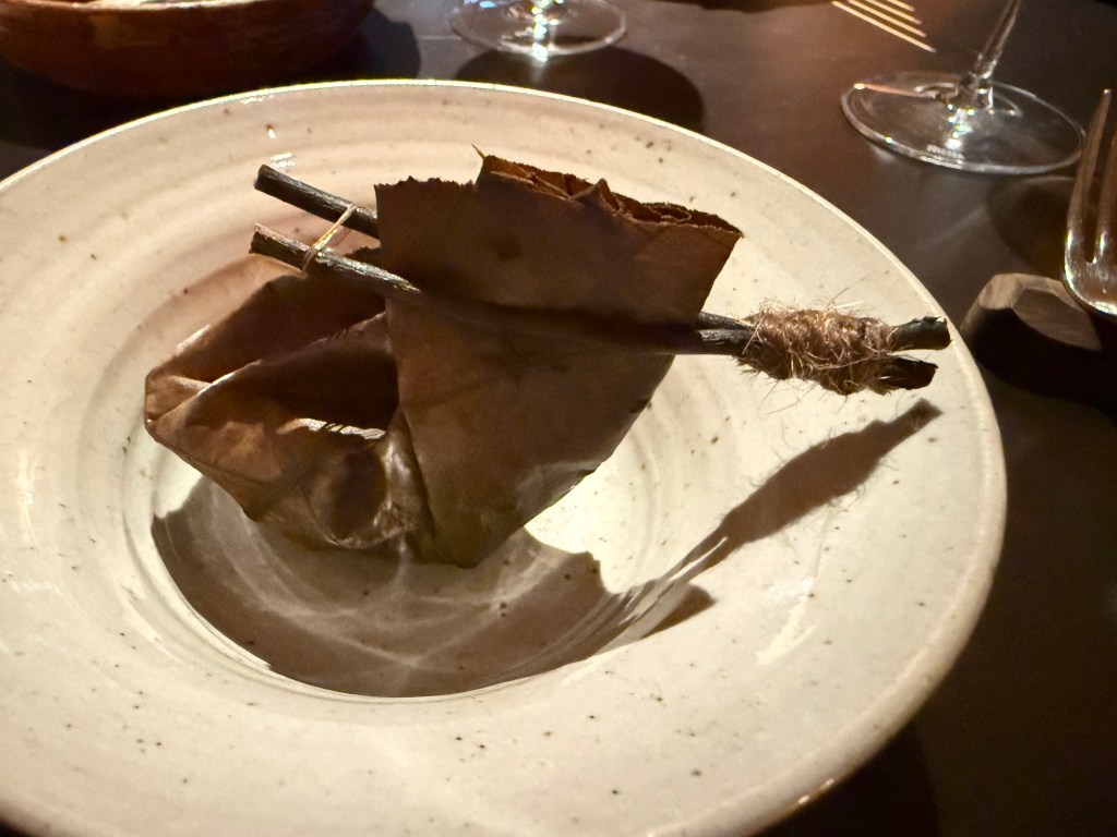 A creatively presented dish featuring a folded leaf resting on a beige plate, secured with twigs and twine.