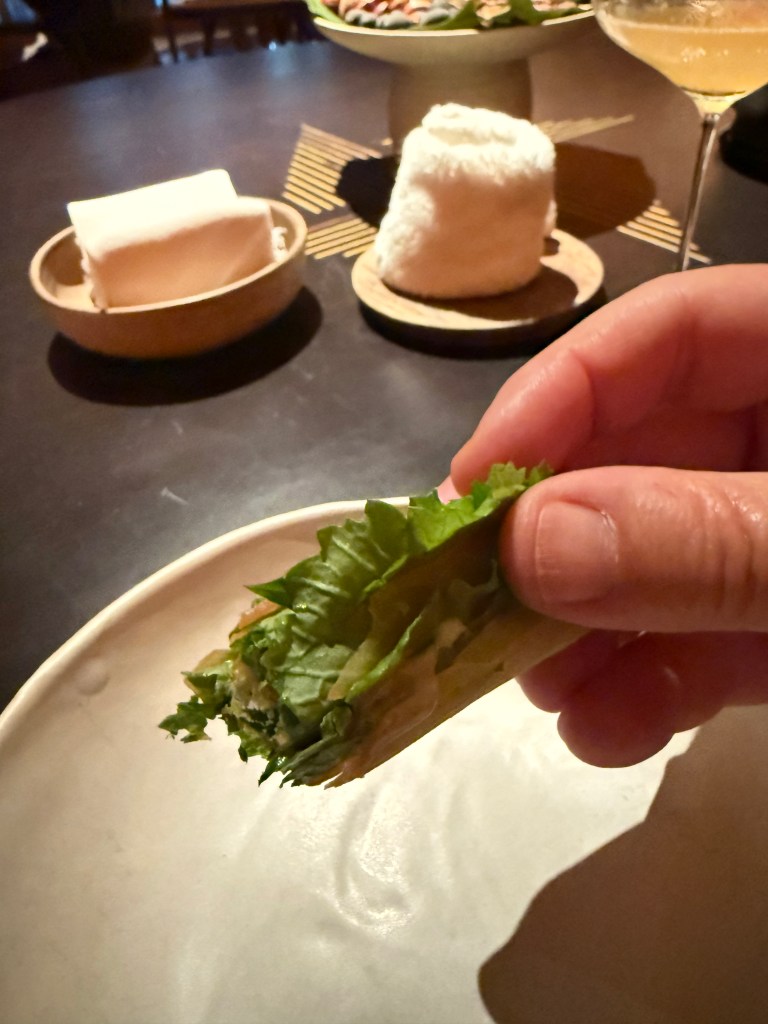 A hand holding a rolled lettuce leaf filled with ingredients, with a plate, sauces, and a drink in the background, set on a dark table.