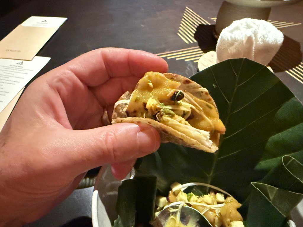A close-up shot of a hand holding a taco filled with various ingredients, set against a dark table with a menu and a white cloth napkin in the background.