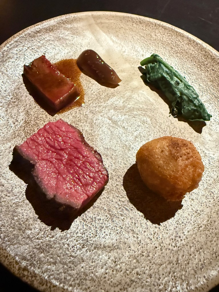 A gourmet dish presented on a textured plate featuring slices of tender beef, a dark sauce, a green vegetable, and a golden fried ball.