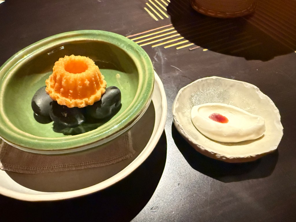 A beautifully plated dessert featuring a small orange cake in a green bowl, surrounded by black stones, alongside a delicate creamy dessert in a white dish.