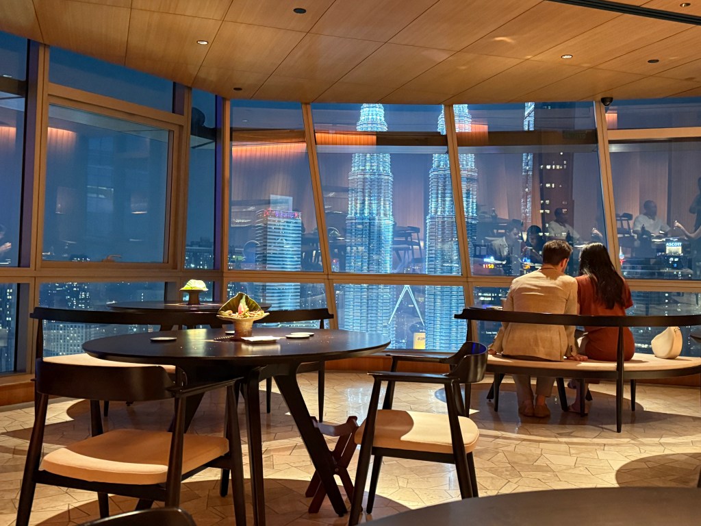 View from inside a restaurant with two people sitting at a table, overlooking the illuminated Petronas Towers at night.
