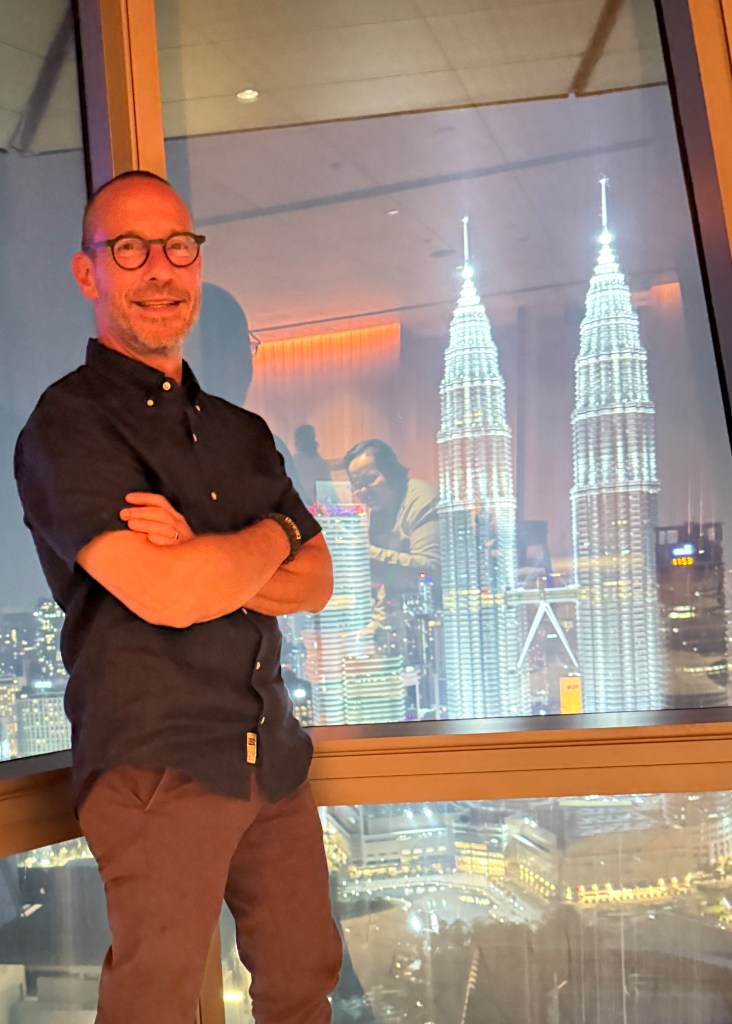 A person standing indoors with their arms crossed, smiling, in front of a large window showcasing the illuminated Petronas Towers at night.