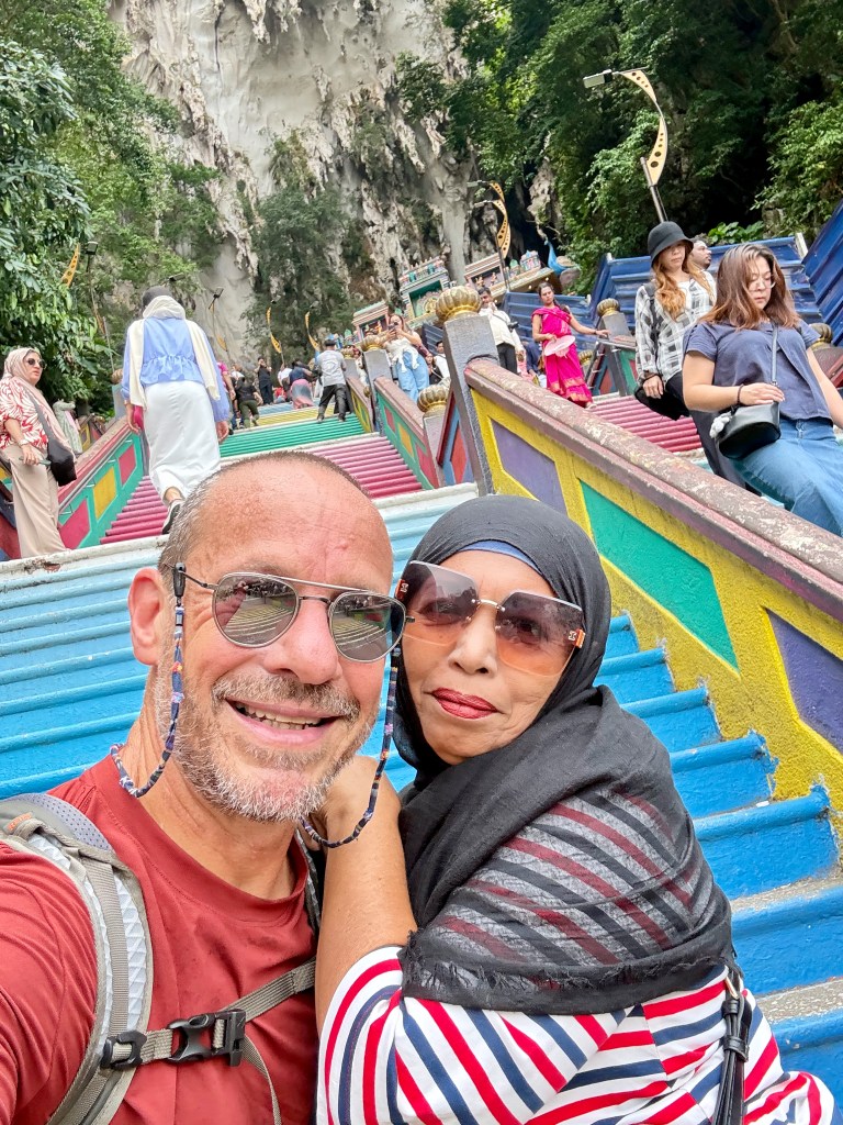 A selfie of two people at the Batu Caves in Malaysia, featuring colorful steps leading up to the cave entrance, with various visitors in the background.