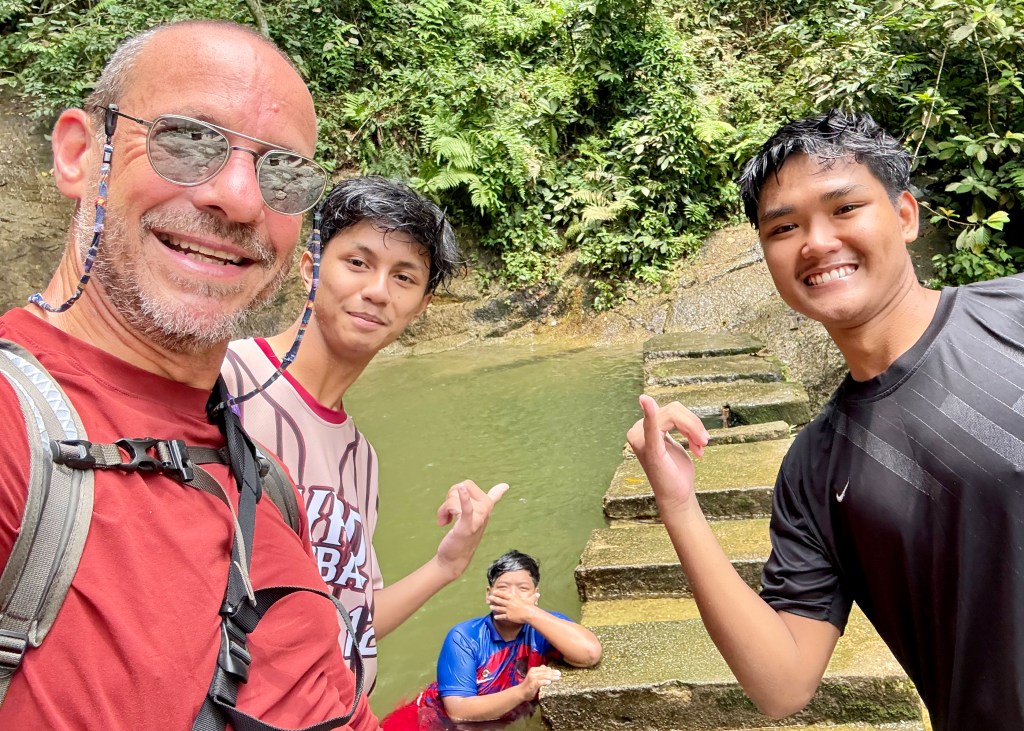 A group of three people smiling for a selfie with natural water pools and lush greenery in the background, at Kanching Waterfalls.