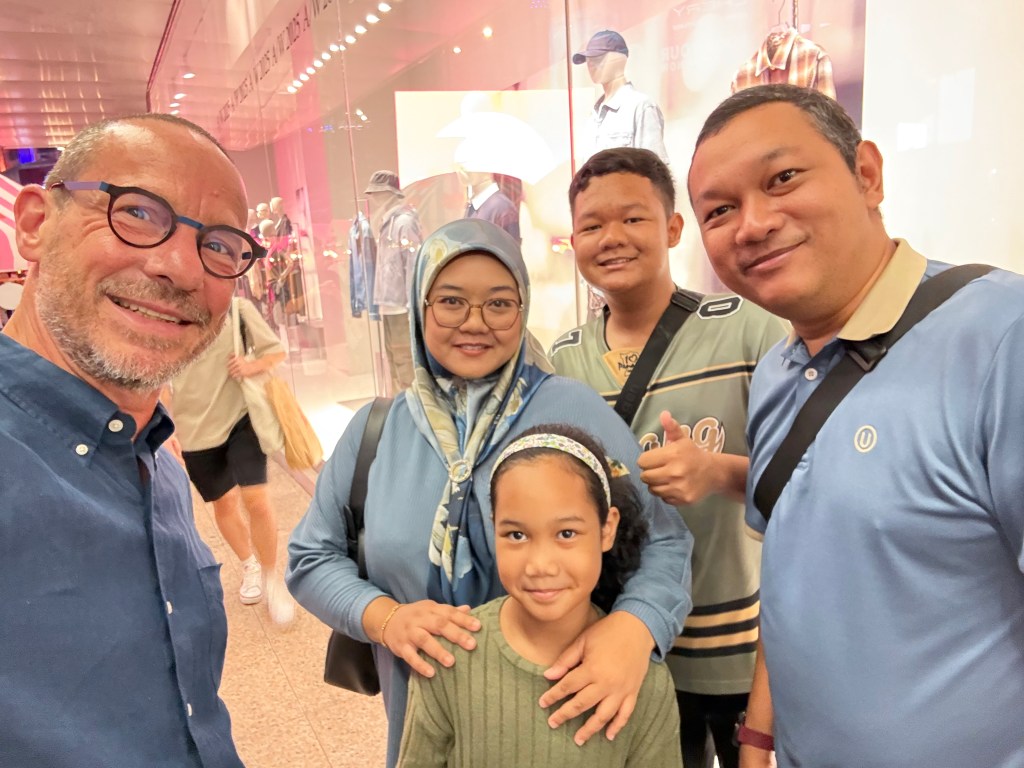 A group selfie featuring a man with glasses and four other individuals, smiling in front of a clothing store display.