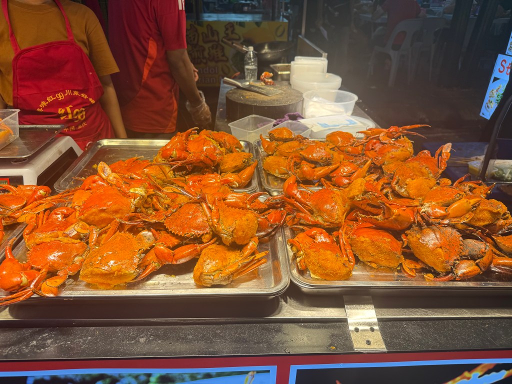 A vibrant food stall displaying a variety of cooked crabs, seasoned and ready to eat, with a vendor in a red apron visible in the background.