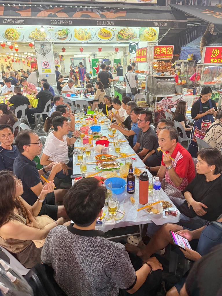A bustling night market scene in Kuala Lumpur with people dining at long tables, enjoying various foods. Food stalls featuring dishes like BBQ stingray and chili crab are visible in the background.