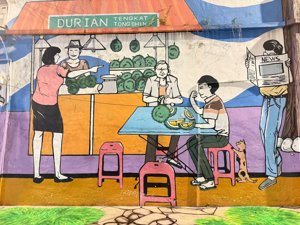 A colorful mural depicting a street scene with individuals enjoying durian, a vendor selling the fruit, and a person reading a newspaper. The artwork features various characters, including a woman talking to a vendor, seated customers, and a dog nearby.