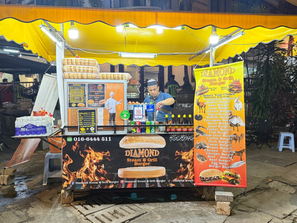 A food stall named 'Diamond Steam & Grill Burger' featuring a variety of burger options, including unique selections like ostrich and horse burgers, with a man preparing food in front of the stall under warm yellow lighting.