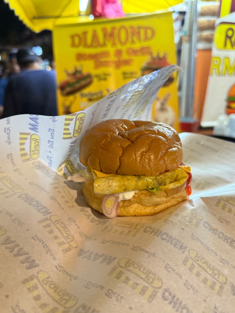 A close-up view of a burger featuring a chicken patty, egg, and various condiments, set against a vibrant food market backdrop.