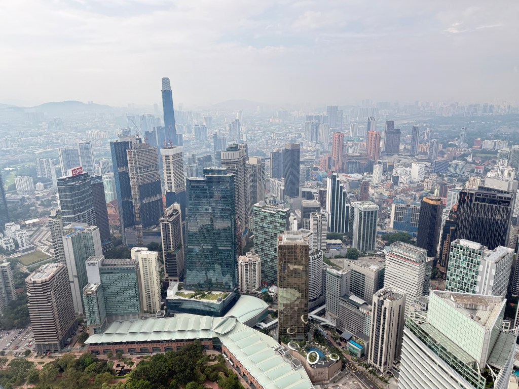 Aerial view of Kuala Lumpur featuring a mix of modern skyscrapers and urban landscape, with notable buildings and greenery visible in the foreground.
