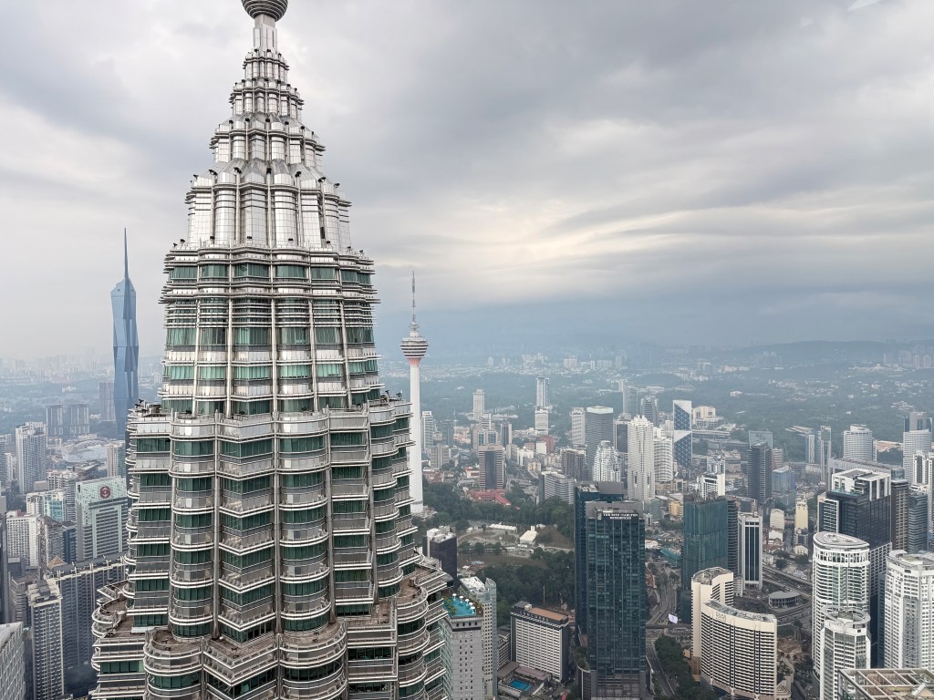 View of the Petronas Towers and surrounding Kuala Lumpur skyline from an observation deck, with cloudy skies.