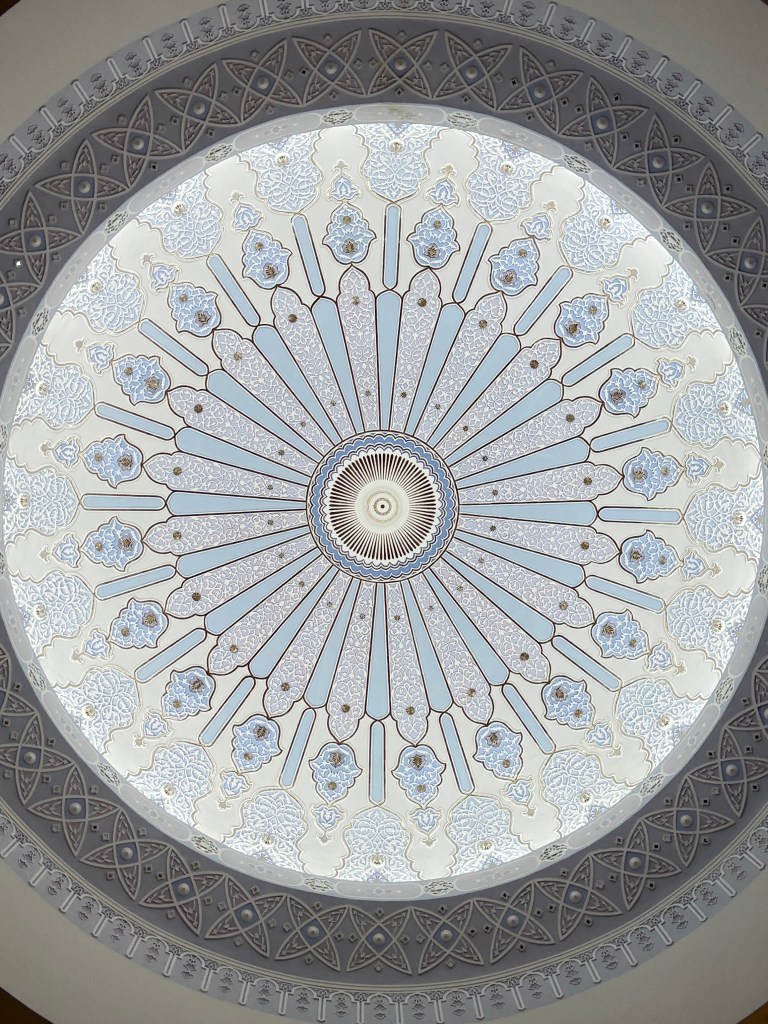 Intricate ceiling design of a mosque with a floral pattern and decorative elements in light blue and cream colors.