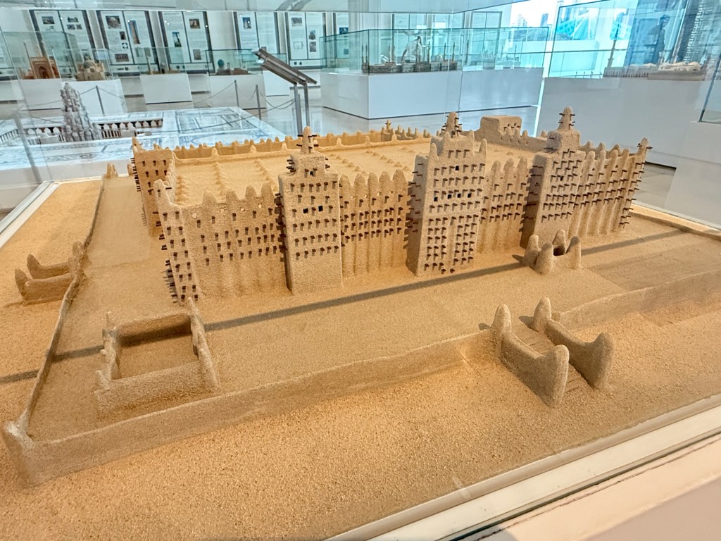 Scale model of a mosque made from sand, showcasing intricate architectural details, displayed in a museum setting.