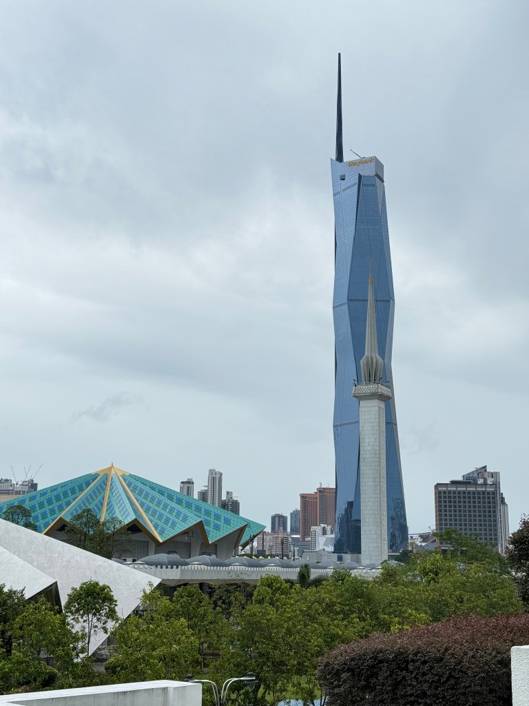 View of the Kuala Lumpur skyline featuring a modern skyscraper with a distinctive design, surrounded by lush greenery and cloudy skies.