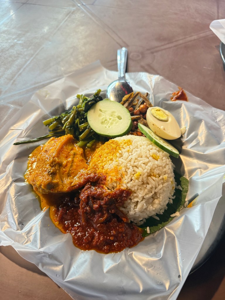 A plate of Nasi Lemak featuring rice, chicken, sambal sauce, fried anchovies, peanuts, cucumber slices, and a hard-boiled egg, served on a banana leaf.