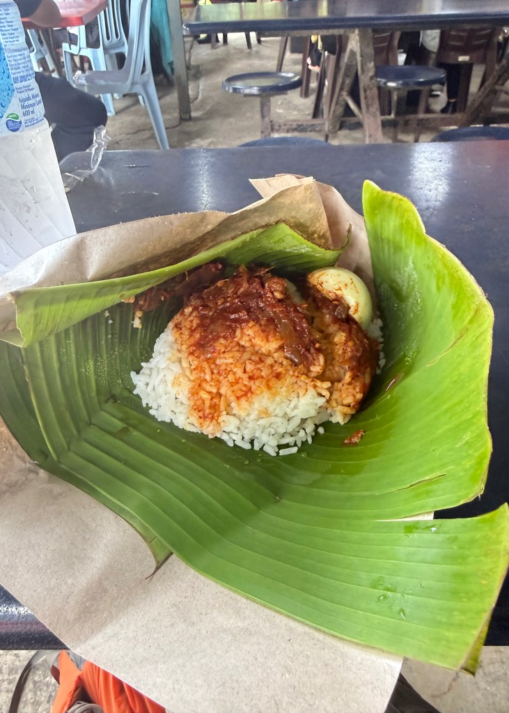 A banana leaf-wrapped package of Nasi Lemak, featuring rice topped with a spicy sauce and accompanied by a boiled egg, set on a table in a casual dining environment.