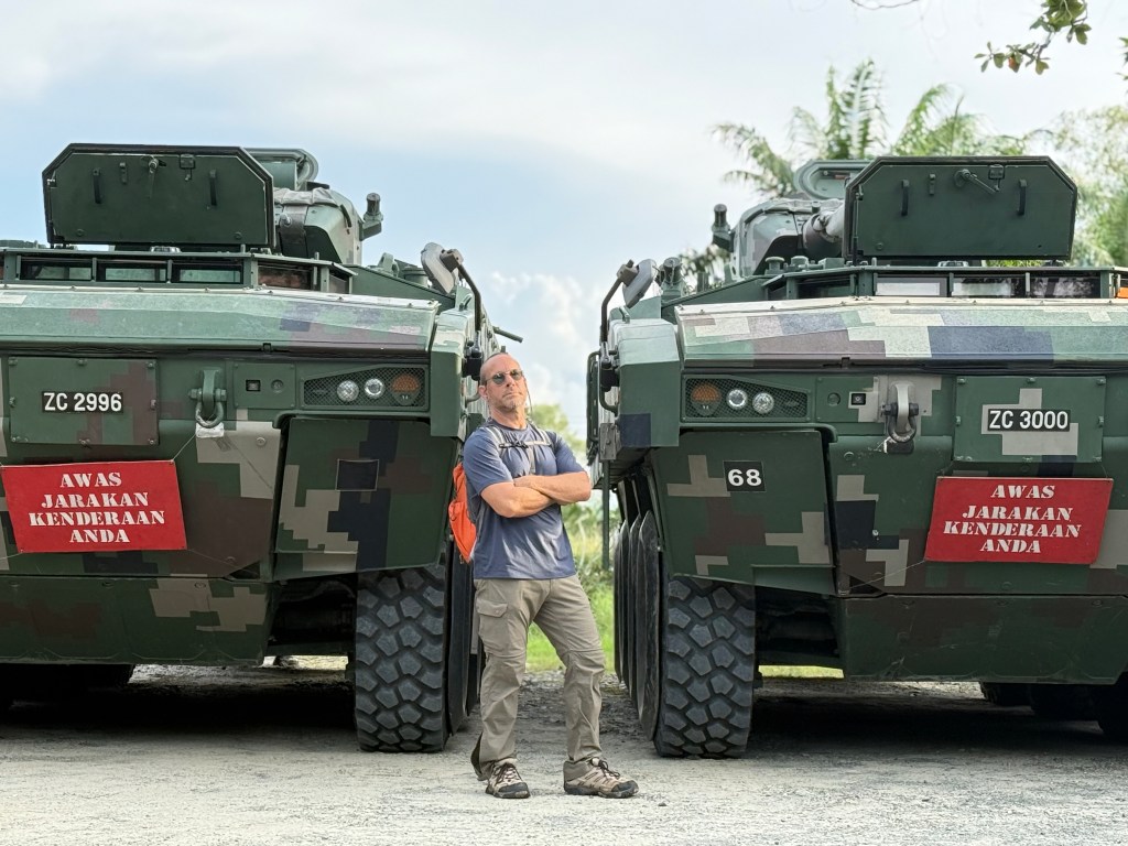 A person standing between two military tanks with warning signs in Malay, outdoors with greenery in the background.