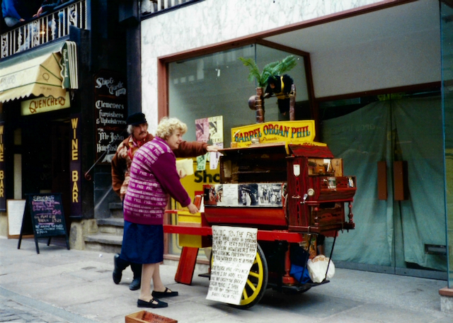A street performer playing a barrel organ named 'Barrel Organ Phil,' with a man interacting with a woman in a pink sweater. The cart features colorful decorations and a small plant, set in an urban area with storefronts in the background.