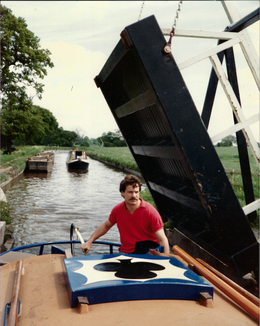 A man in a red shirt navigates a boat along a canal with a raised bridge in the background. Two other boats are visible on the water, surrounded by green trees.