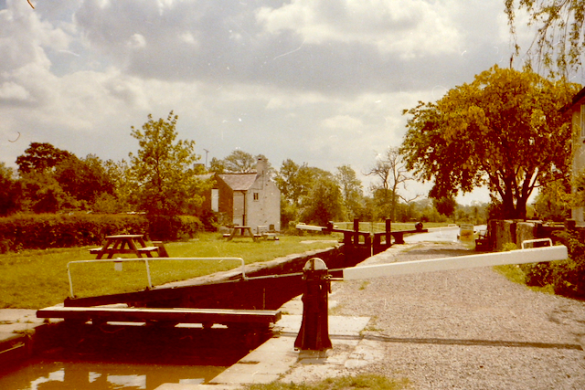 A scenic view of a canal lock system with wooden gates, surrounded by lush greenery and a quaint stone house. A path runs alongside the canal, with picnic tables visible in the foreground.