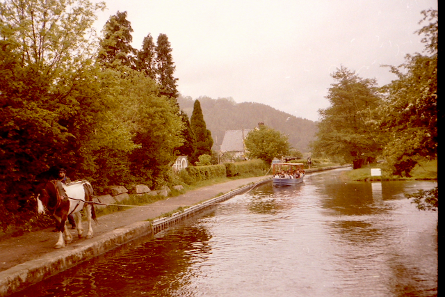 A canal scene featuring a horse pulling a barge alongside a lush, green bank. Trees and a distant house are visible, along with a boat carrying passengers on the calm water.