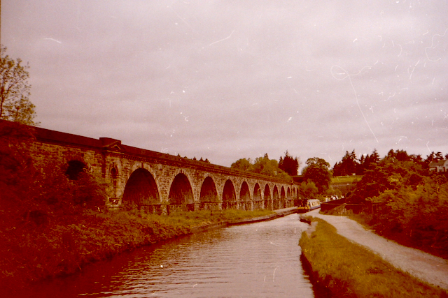 A historical stone railway viaduct with multiple arches stretching over a river, surrounded by greenery under a cloudy sky.