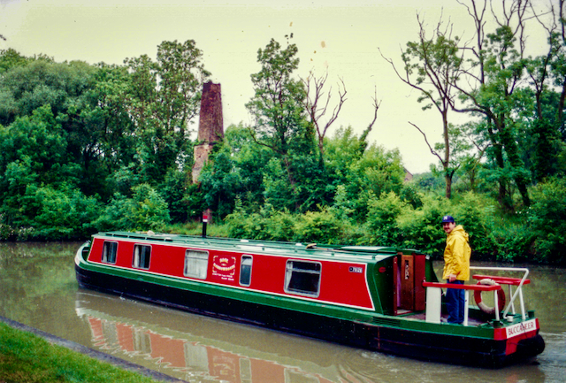 A colorful canal boat with a man wearing a yellow raincoat, navigating through a lush green landscape along a waterway, with a ruined tower visible in the background.
